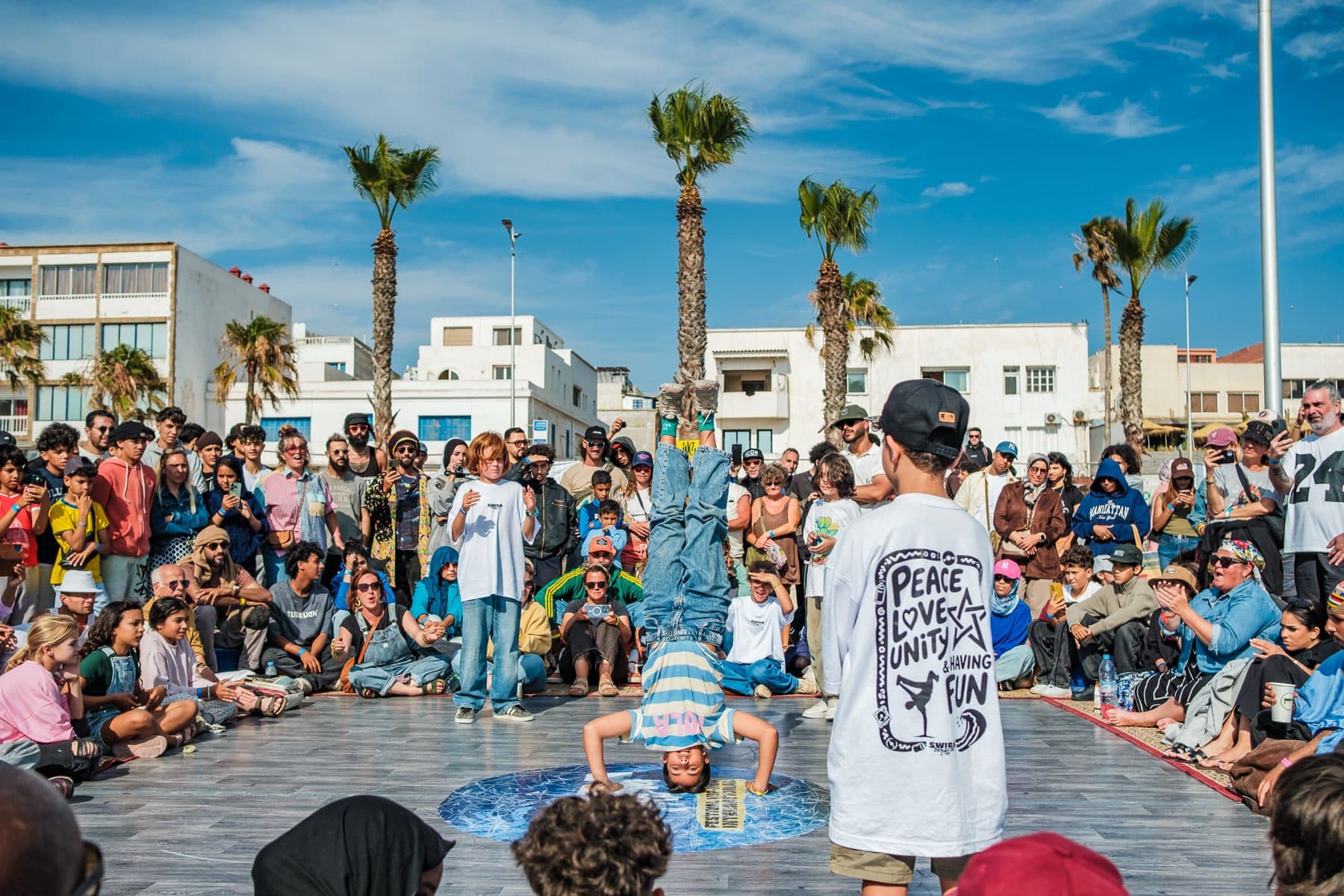 Un enfant en équilibre sur la tête au centre du cercle, entouré du public, sur la corniche d'Essaouira.